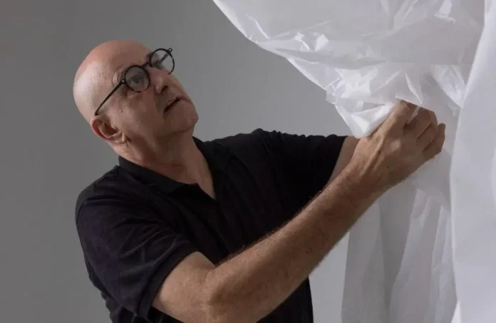 Homem careca usando óculos ajusta um tecido branco em um fundo cinza.