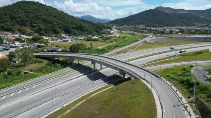 Vista aérea de um viaduto sobre uma rodovia, cercado por áreas verdes e construções.