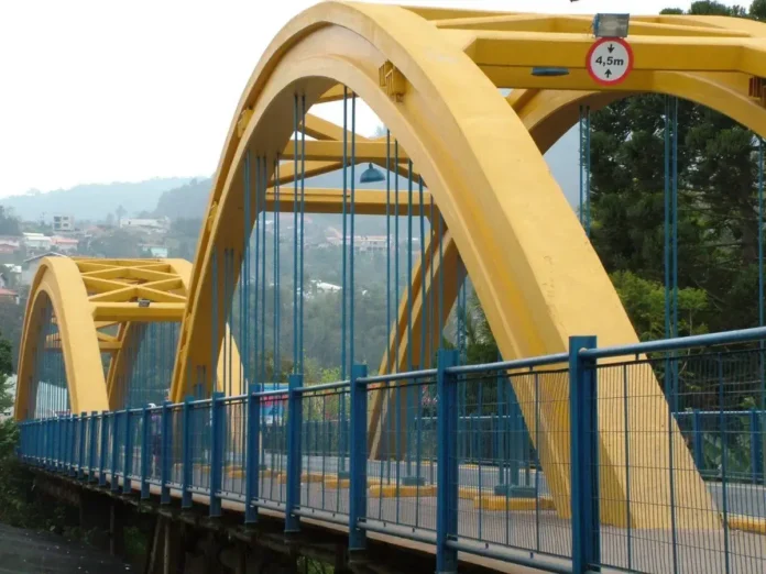 Ponte amarela com arcos e estrutura metálica, cercada por árvores e edificações ao fundo. Sinalização de altura visível.