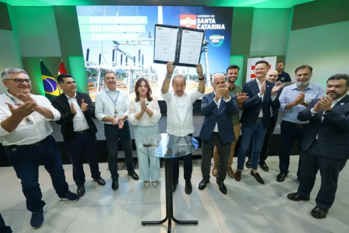 Grupo de pessoas aplaudindo enquanto um homem exibe documentos em um evento do Programa Energia Boa, com fundo ilustrativo de infraestrutura elétrica.