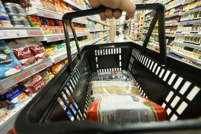 Cesta de compras visível com produtos como arroz e farinha, em um supermercado com prateleiras cheias.
