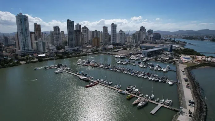 Vista aérea de um porto com vários barcos e arranha-céus ao fundo, com céu claro e nuvens.