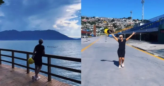 Pessoa vestindo roupa preta e boné, em um deck de madeira, olhando para o mar e montanhas ao fundo.