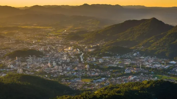 Vista panorâmica de uma cidade cercada por montanhas, com edifícios e vegetação visíveis ao entardecer.