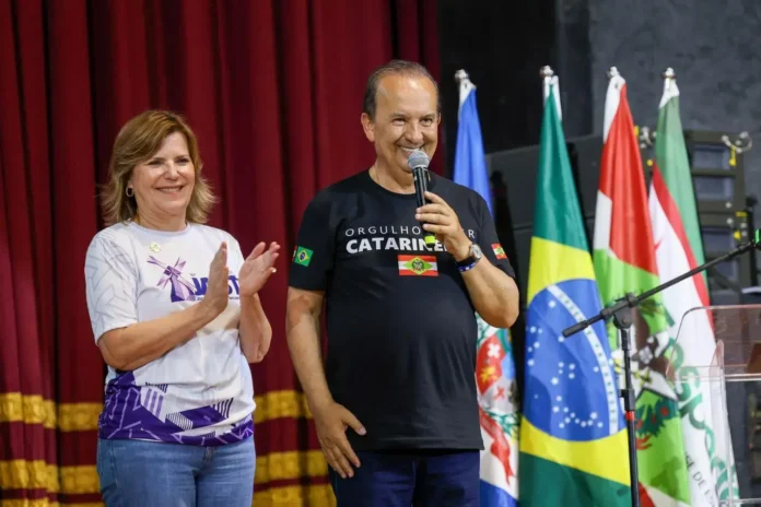 Duas pessoas em um palco, uma aplaudindo e a outra segurando um microfone, com bandeiras ao fundo.