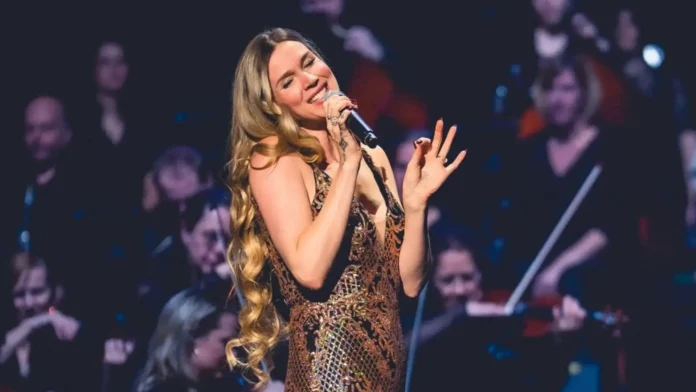 Cantora em performance ao vivo, segurando um microfone, com público e músicos ao fundo.