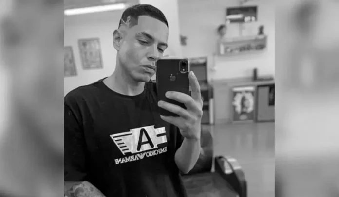 Homem segurando um celular, com cabelo estilizado e vestindo camiseta preta em ambiente interno.