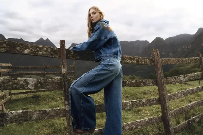 Pessoa usando um conjunto de jeans, encostada em uma cerca de madeira, com montanhas ao fundo.