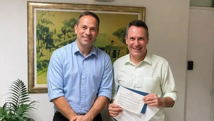 Dois homens sorrindo em um ambiente interno, segurando documentos. Ao fundo, uma parede com um quadro decorativo.