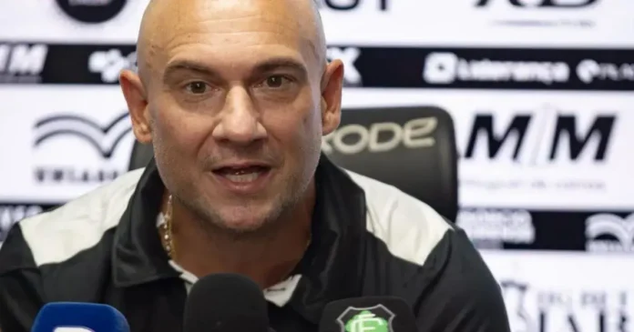 Treinador em coletiva, expressando-se e cercado por microfones, com painel de fundo e logo do clube.