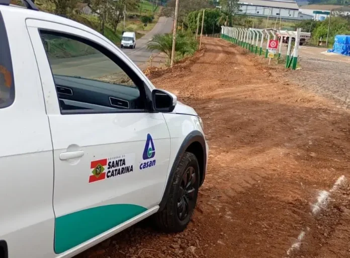 Veículo da Casan estacionado em estrada de terra, com vegetação ao fundo e edificações distantes.