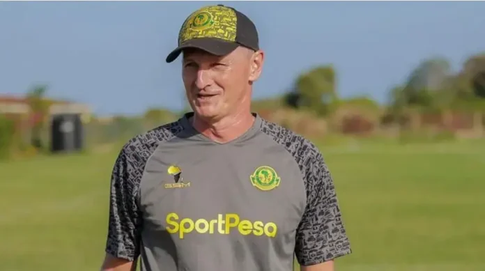 Treinador sorrindo, vestindo camiseta cinza com estampa e boné, em campo com grama ao fundo.