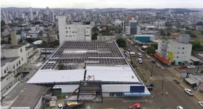 Vista aérea de um prédio em reforma, com telhado parcialmente coberto, cercado por ruas e outros edifícios ao fundo.