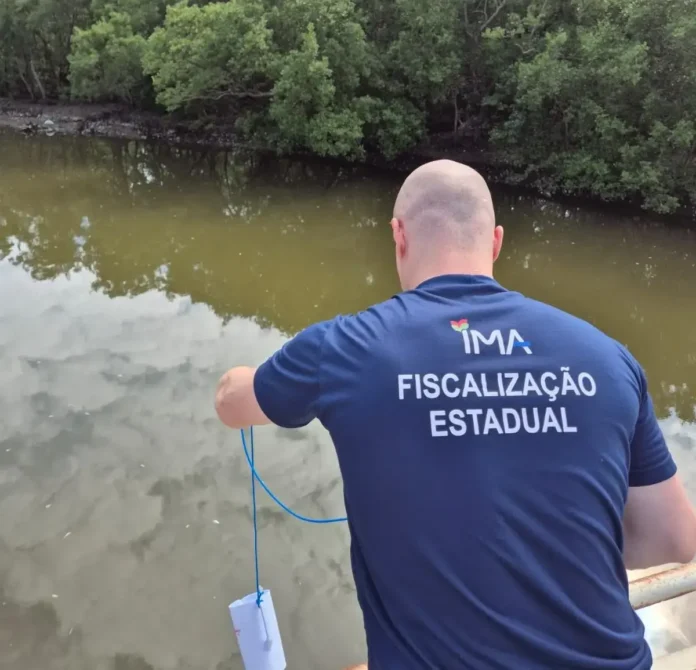 Pessoa realizando amostragem em corpo d'água, com vegetação ao fundo e refletindo a superfície. Texto visível: "FISCALIZAÇÃO ESTADUAL".