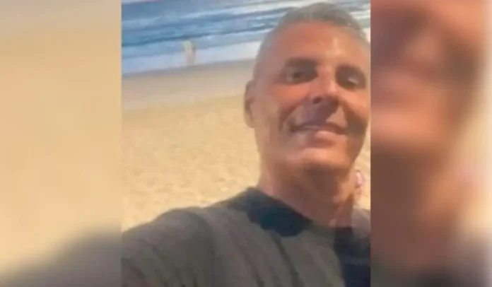 Selfie de um homem na praia, com ondas e areia ao fundo.