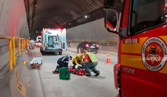 Equipes de socorro atendem vítima em túnel, com ambulância e viatura do Corpo de Bombeiros Militar ao fundo.
