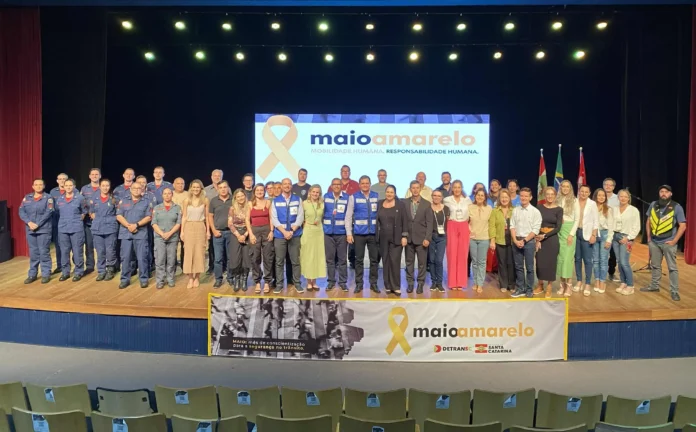 Grupo de pessoas posando em palanque, com banner do Maio Amarelo e mensagem sobre mobilidade e responsabilidade humana.