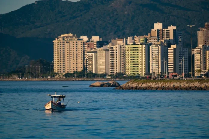 Barco pequeno navegando em águas calmas com edifícios altos ao fundo e montanhas.