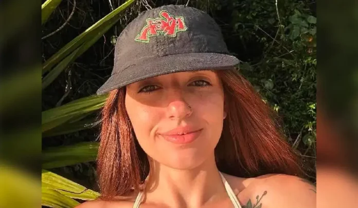 Pessoa sorrindo, com cabelo longo e castanho, usando um chapéu escuro, em meio a vegetação.