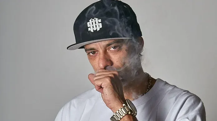 Homem com boné preto e camiseta branca, respirando fumaça e olhando para a câmera.