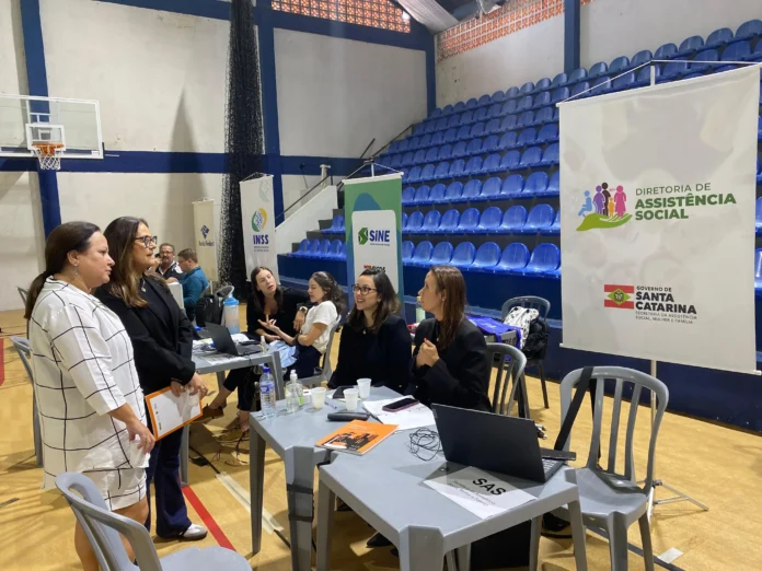 Grupo de profissionais em um evento, conversando e realizando atendimentos em uma quadra esportiva. Banners da Diretoria de Assistência Social visíveis.