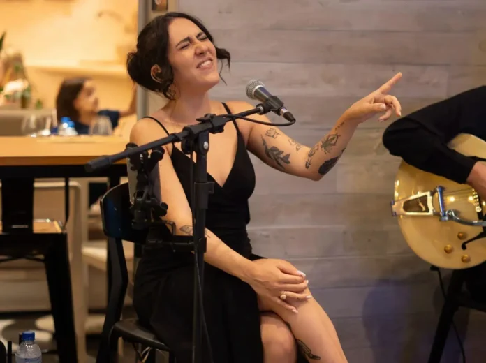 Cantora performando em microfone, expressando emoção enquanto senta em uma cadeira, acompanhada por músico ao fundo.