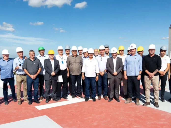 Grupo de pessoas usando capacetes de segurança, posando em uma obra, com céu claro ao fundo.