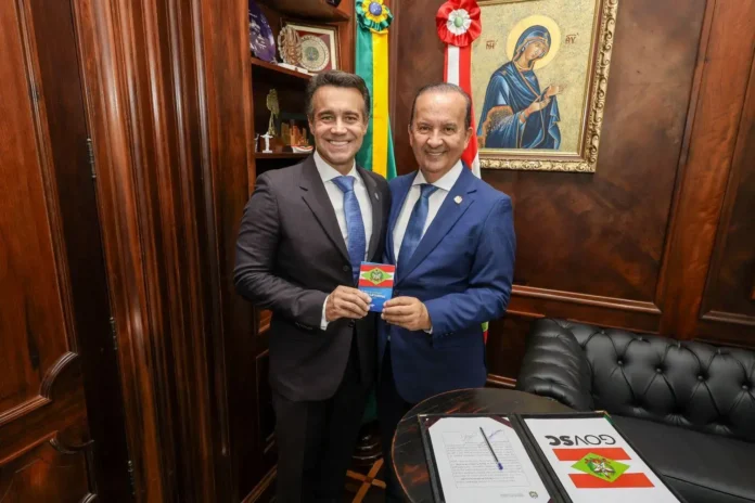 Duas pessoas sorrindo em um escritório, segurando um documento com bandeira e brasão, ao fundo um quadro religioso.