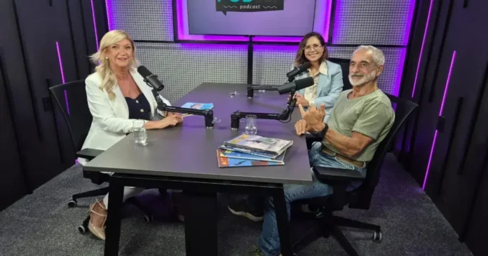 Três pessoas sentadas em uma mesa de podcast, com microfones e materiais de leitura ao fundo.