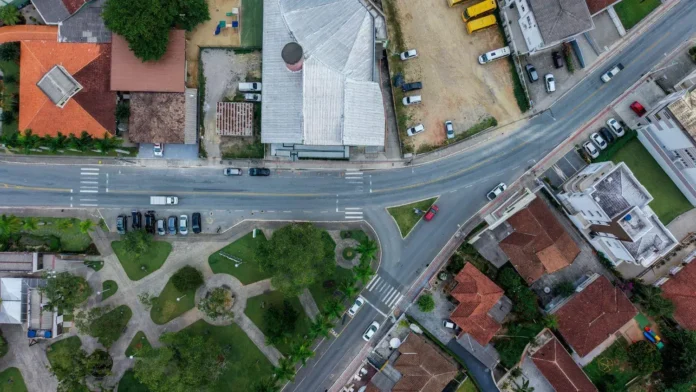 Vista aérea de um cruzamento em uma área urbana, com edifícios, ruas, veículos estacionados e áreas verdes.