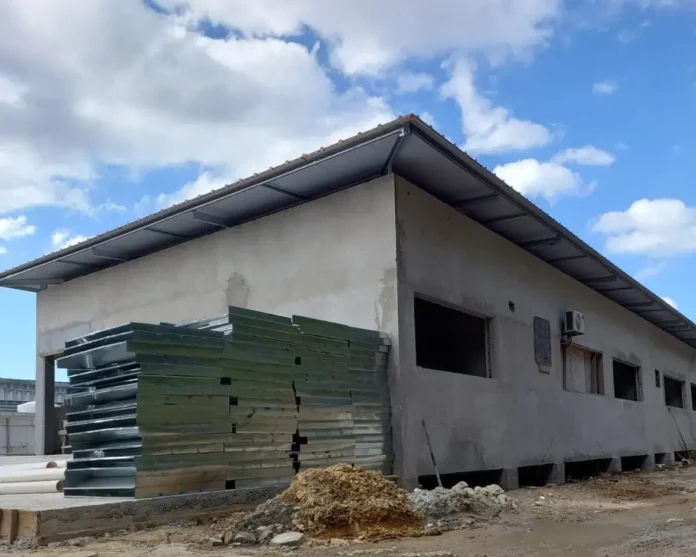 Construção em andamento com paredes externas sem acabamento, materiais e entulho ao redor, céu com nuvens.