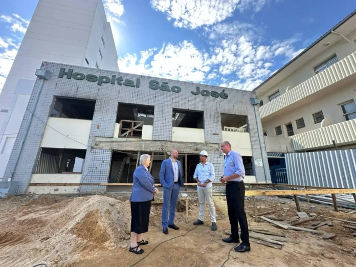 Quatro pessoas conversam em frente à obra do Hospital São José, com parte da construção em andamento e céu azul ao fundo.