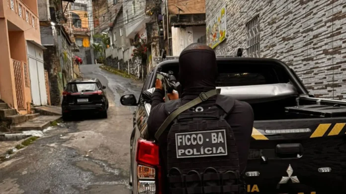 Policial armado com farda tática aponta arma para uma rua em área urbana. Veículo de polícia estacionado ao fundo.