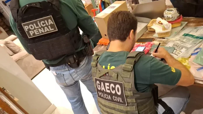 Agentes de polícia com coletes identificados da Polícia Penal e GAECO em ação, realizando investigações em um ambiente interno.