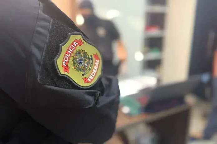 Braço de agente da Polícia Federal com o distintivo visível, em ambiente interno de operação.
