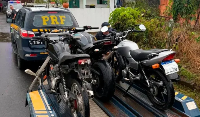 Veículos apreendidos em um reboque com viatura da polícia ao fundo em uma via.