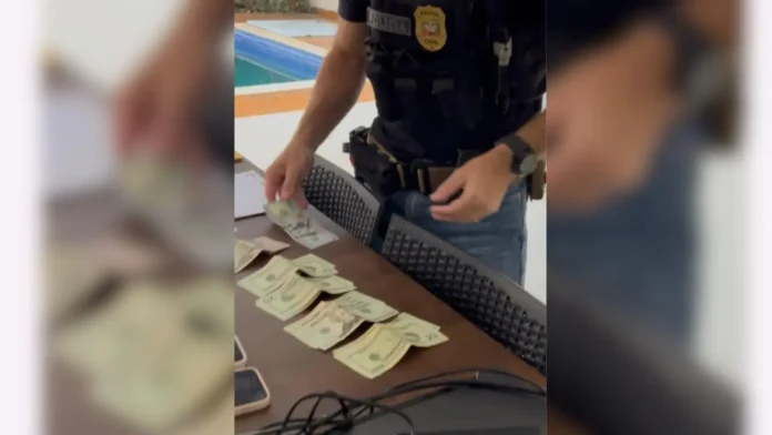 Pessoa organizando notas de dinheiro sobre uma mesa, com um celular e um computador ao fundo.