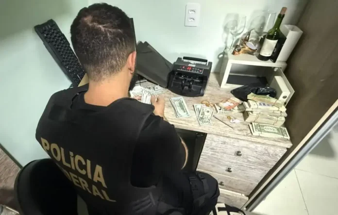Homem em colete da polícia contando dinheiro em uma mesa com notas e maquininha de contagem.