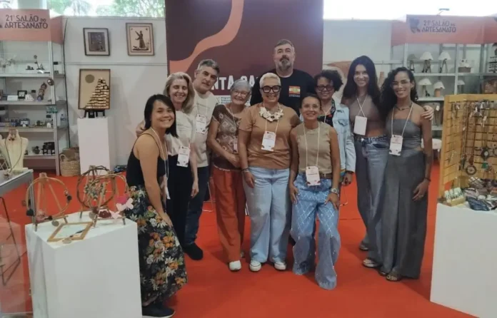 Grupo de pessoas sorrindo em um estande no 21º Salão do Artesanato, com produtos artesanais exibidos ao fundo.