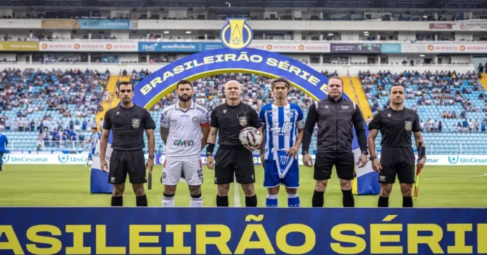 Jogadores e árbitros posam frente ao arco "Brasileirão Série B" em campo. Torcedores ao fundo.
