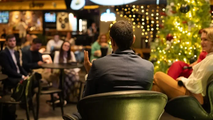 Grupo de pessoas assistindo a uma apresentação em ambiente descontraído, decorado com luzes e uma árvore de Natal ao fundo.