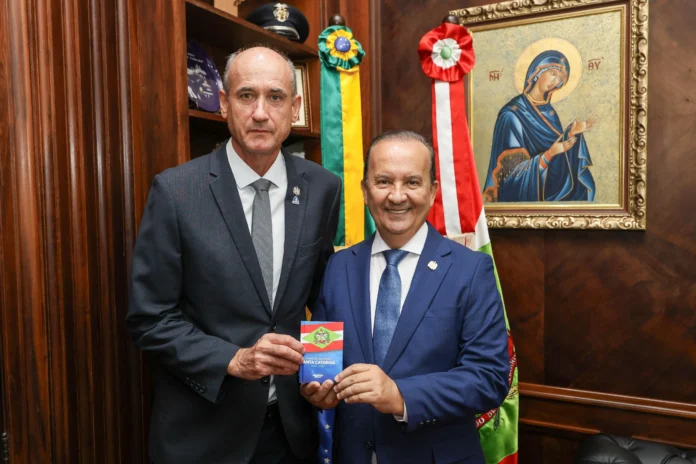 Duas pessoas em um ambiente interno segurando um documento com bandeira, com bandeiras e uma imagem religiosa ao fundo.