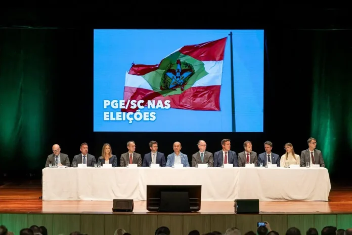 Painel com representantes em evento sobre eleições, ao fundo uma tela com a frase "PGE/SC NAS ELEIÇÕES" e bandeira.