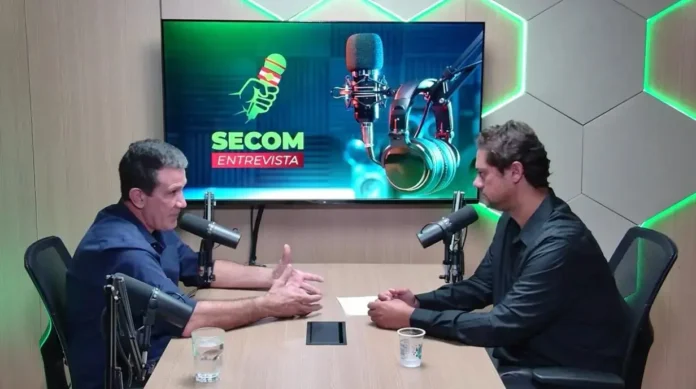 Dois homens em uma mesa discutindo, com microfones e um fundo que exibe o logotipo do podcast "Secom Entrevista".