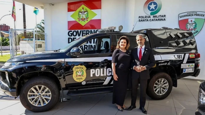 Pessoa e homem em trajes formais ao lado de veículo policial, com o logotipo da Polícia Civil de Santa Catarina.