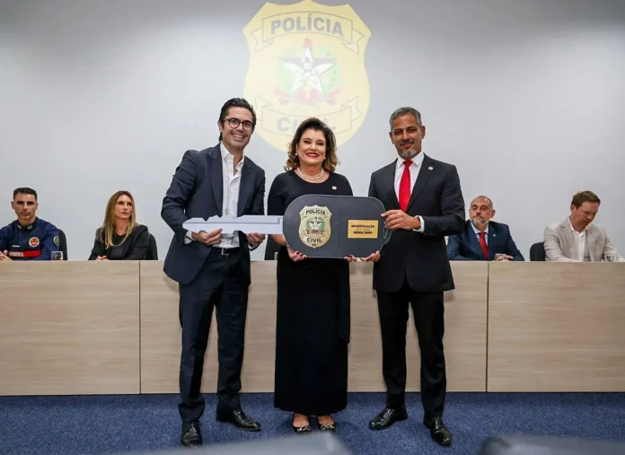 Três pessoas seguram uma chave grande com o símbolo da Polícia Civil, em evento com autoridades ao fundo.