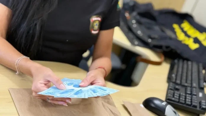 Mãos segurando várias cédulas azuis sobre uma mesa com teclado e objetos ao fundo.
