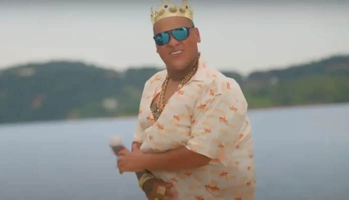 Homem usando uma camisa com estampa de peixes, correntes de metal, óculos escuros e uma coroa, posando em frente a um fundo de lago.