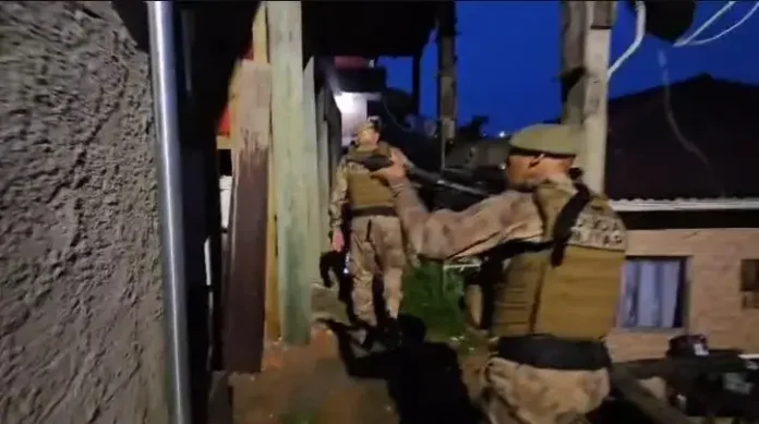 Militares armados em operação noturna, caminhando por uma rua estreita entre casas, em ambiente urbano.