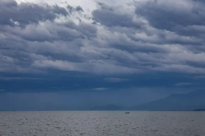 Céu nublado e mar calmo com um pequeno barco à deriva, ao fundo montanhas distantes.